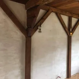 Drewniana konstrukcja altany z belkami i lampą naftową w stylu retro, ściany wykończone tynkiem strukturalnym.