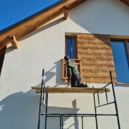 Pracownik na rusztowaniu montuje drewnopodobną okładzinę elewacyjną wokół okna w domu jednorodzinnym.