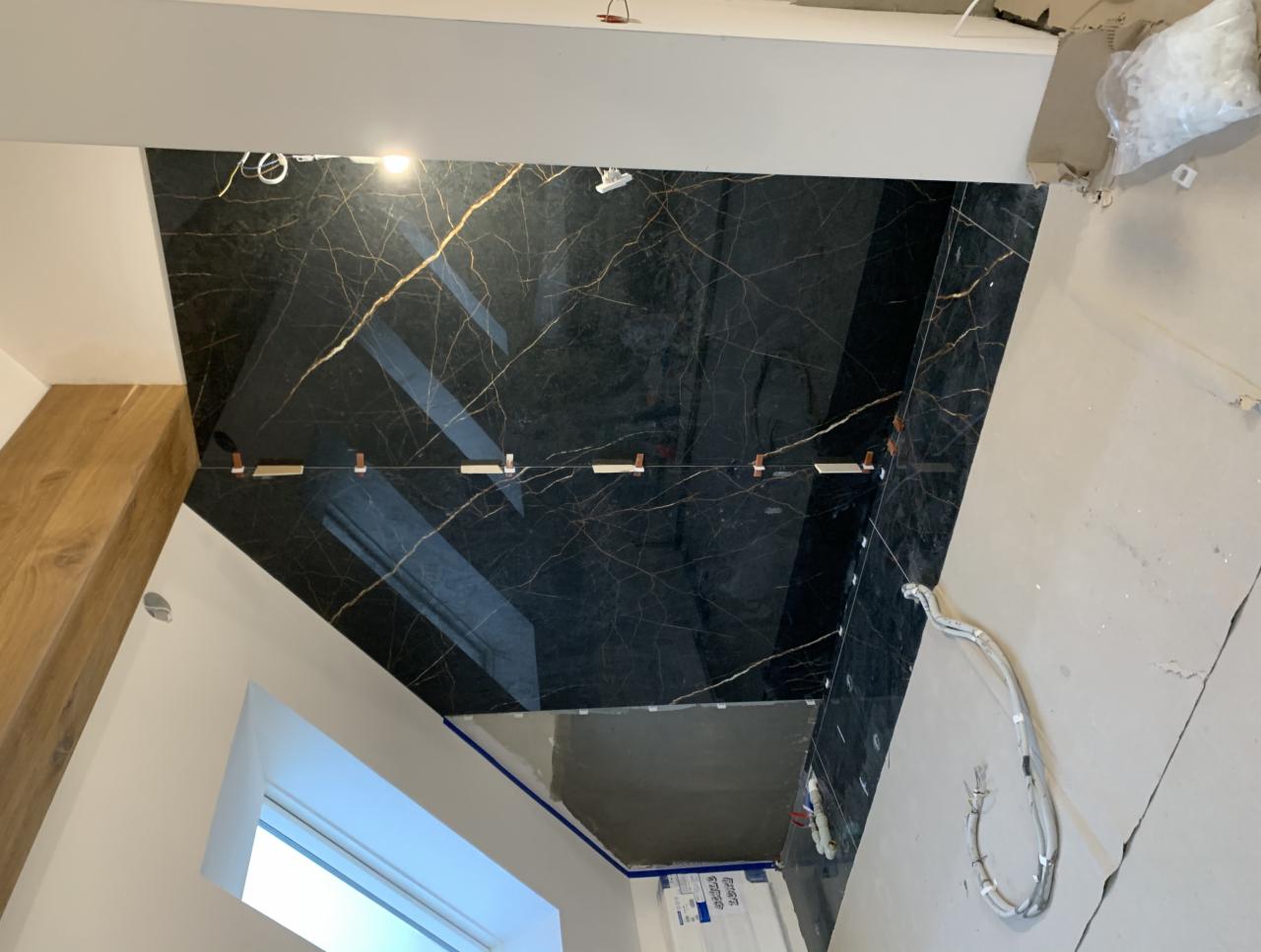 Montaż czarnych płytek imitujących marmur z wyraźnym, złotym żyłowaniem na ścianie, widoczne krzyżyki dystansowe, przewody elektryczne i okno dachowe w trakcie prac wykończeniowych.