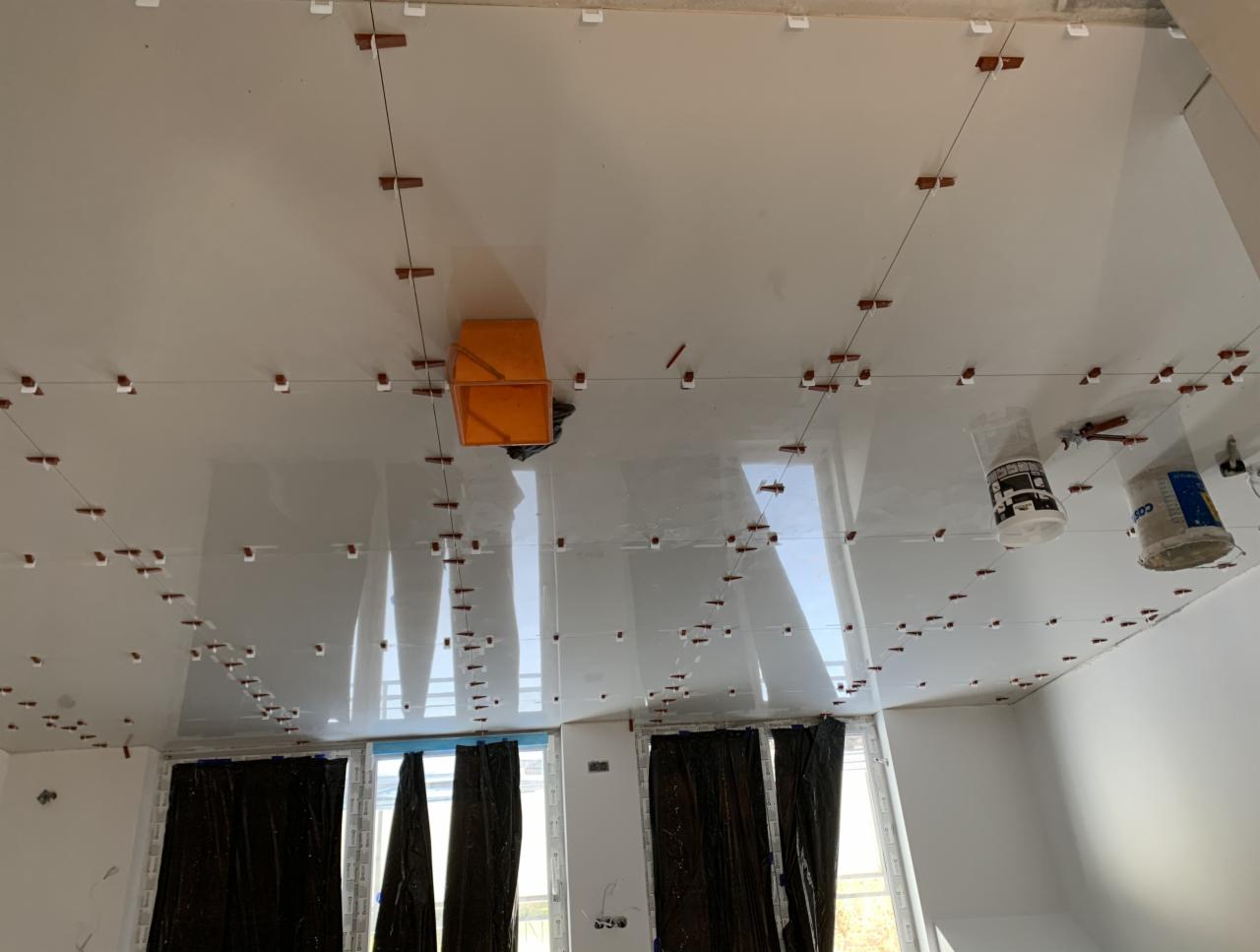 Montaż płyt kartonowo-gipsowych na suficie z użyciem sznurków i poziomicy, widoczne akcesoria montażowe i okna zasłonięte folią.
