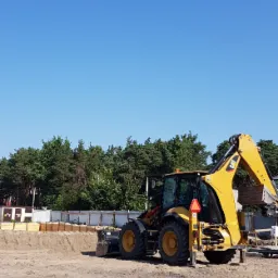 Żółta koparko-ładowarka CAT na placu budowy z widocznym wykopem pod fundamenty i szalunkiem drewnianym, las w tle i błękitne niebo