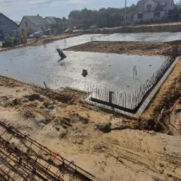 Świeżo wylany fundament z wystającymi prętami zbrojeniowymi, otoczony piaskiem i wykopem. W tle widoczne domy i sprzęt budowlany.