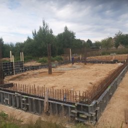 DOK-BUD Krystian Szukalski - Zbrojenie fundamentów domu jednorodzinnego w szalunkach systemowych, widoczne wystające pręty zbrojeniowe, teren budowy, pochmurne niebo.