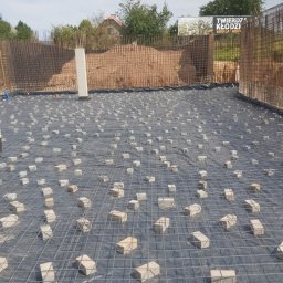 DOK-BUD Krystian Szukalski - Zbrojenie fundamentu z siatki stalowej na czarnej folii, podparte kostkami. W tle pionowe zbrojenie ścian fundamentowych i częściowo widoczny pracownik w czerwonej rękawicy.