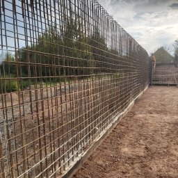 DOK-BUD Krystian Szukalski - Siatka zbrojeniowa pionowa, przygotowana do zalania betonem, widoczna z perspektywy ziemi, z pracownikami w tle i elementami szalunku.