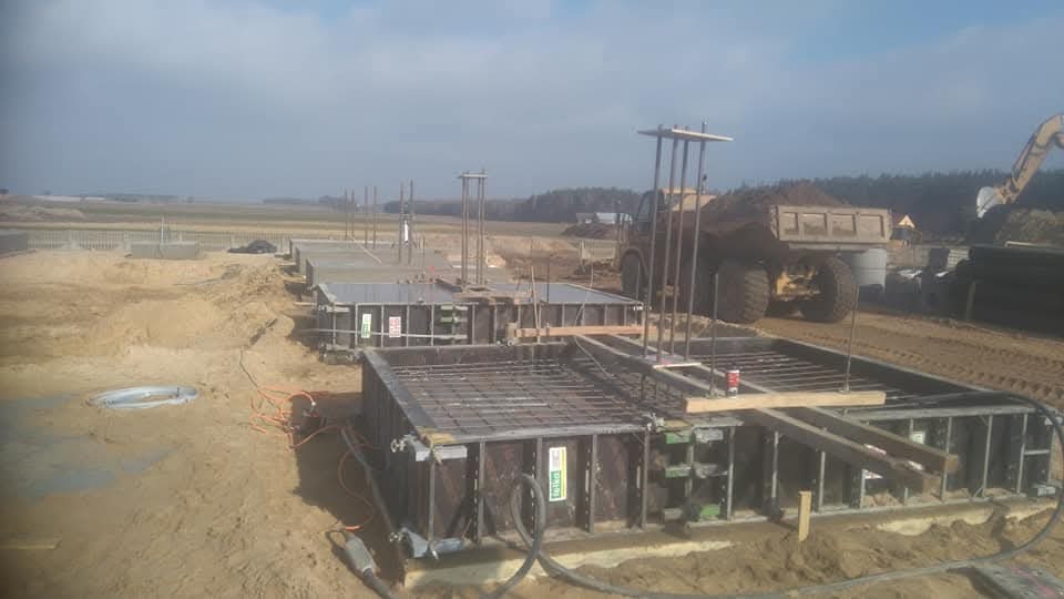 Przygotowane szalunki i zbrojenie pod fundamenty, widoczny ciężki sprzęt budowlany w tle.