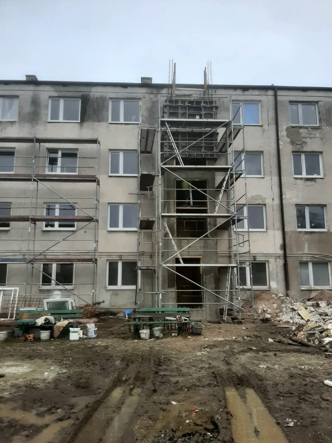 Renowacja elewacji budynku wielorodzinnego z użyciem rusztowań, widoczne elementy zbrojenia na szczycie, błotniste podłoże i gruz.