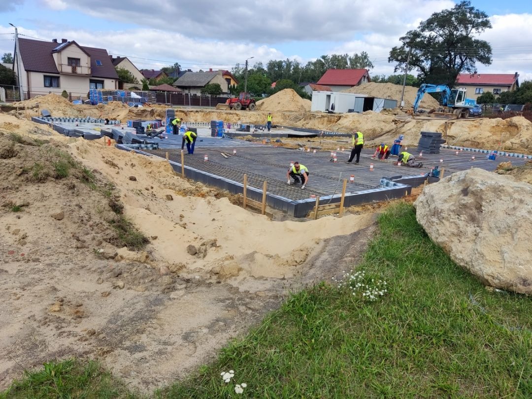 Prace zbrojarskie przy fundamentach domu jednorodzinnego. Widoczni pracownicy w żółtych kamizelkach, zbrojenie i przygotowany teren pod budowę.