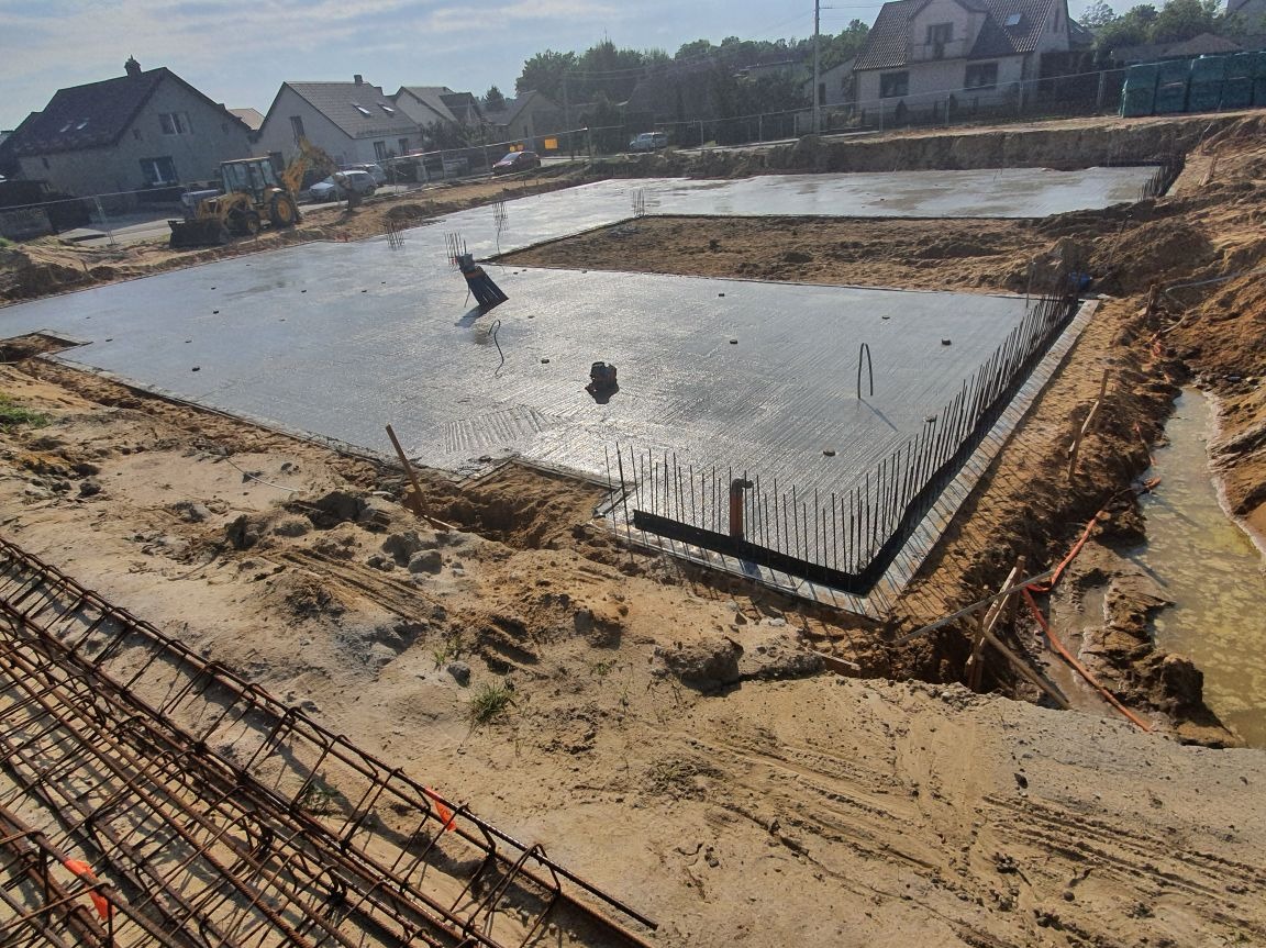 Świeżo wylany fundament z wystającymi prętami zbrojeniowymi, otoczony piaskiem i wykopem. W tle widoczne domy i sprzęt budowlany.