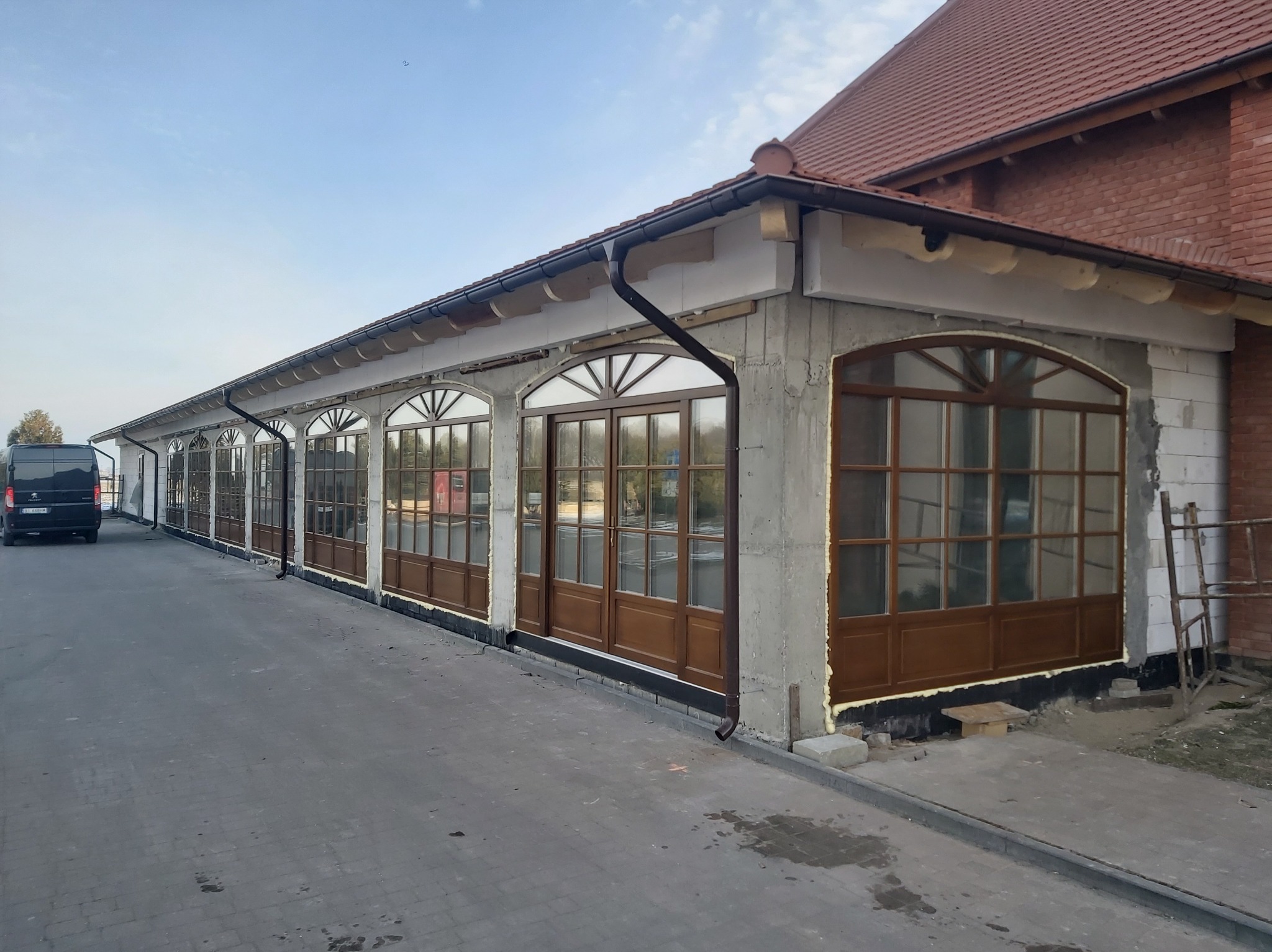 Budynek w trakcie prac wykończeniowych z szeregiem okien z brązowymi ramami. Widoczna elewacja w stanie surowym i fragment dachu z czerwonej dachówki.