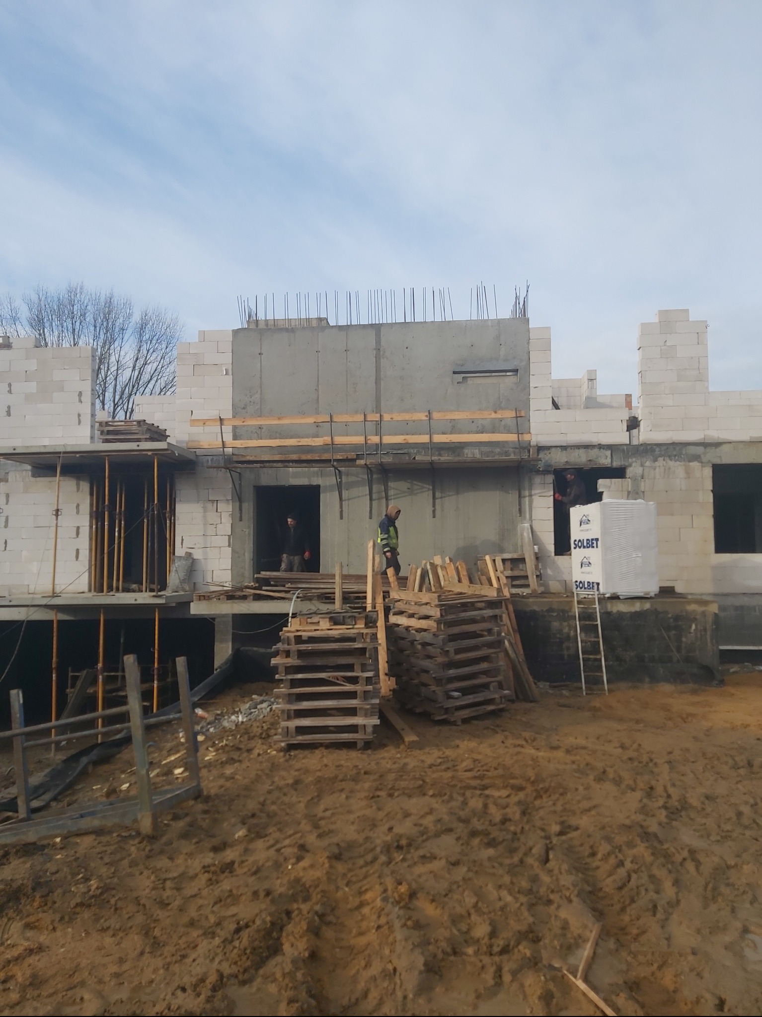 Wznoszenie ścian domu z betonu komórkowego, widoczne zbrojenia i palety z materiałami budowlanymi. Na pierwszym planie ziemia i błoto.