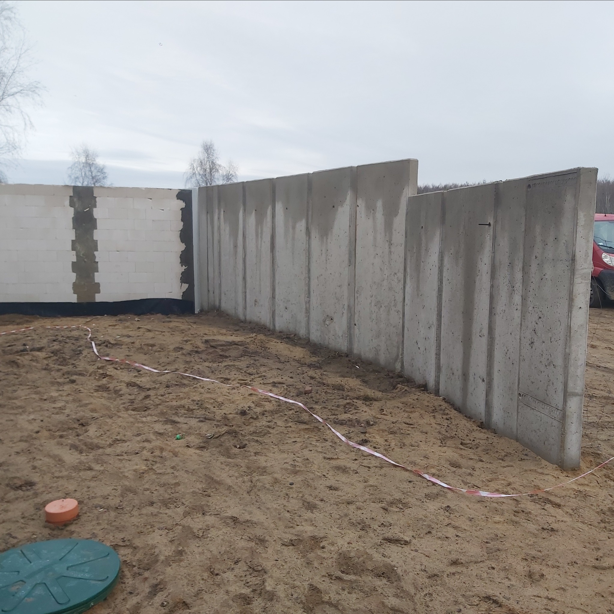 Wznoszenie ścian z betonu i bloczków na budowie. Widoczne szalunki i fragmenty muru. Prace budowlane w toku, teren przygotowany, widoczne oznaczenia.