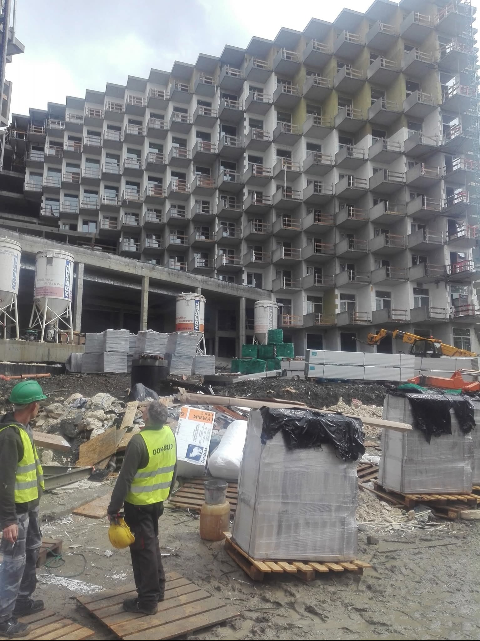 Wielopiętrowy budynek w trakcie budowy z charakterystycznymi balkonami; na pierwszym planie robotnicy budowlani i materiały budowlane.