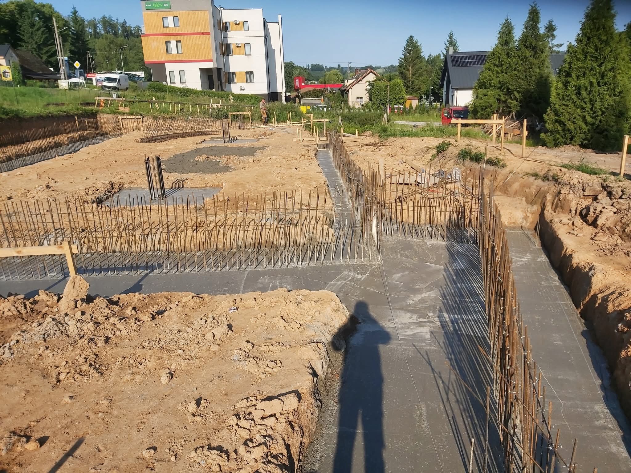 Zalane fundamenty z wystającą zbrojeniem na placu budowy. W tle budynek mieszkalny i zieleń. Cień osoby w kadrze. Widok z góry na wykop.