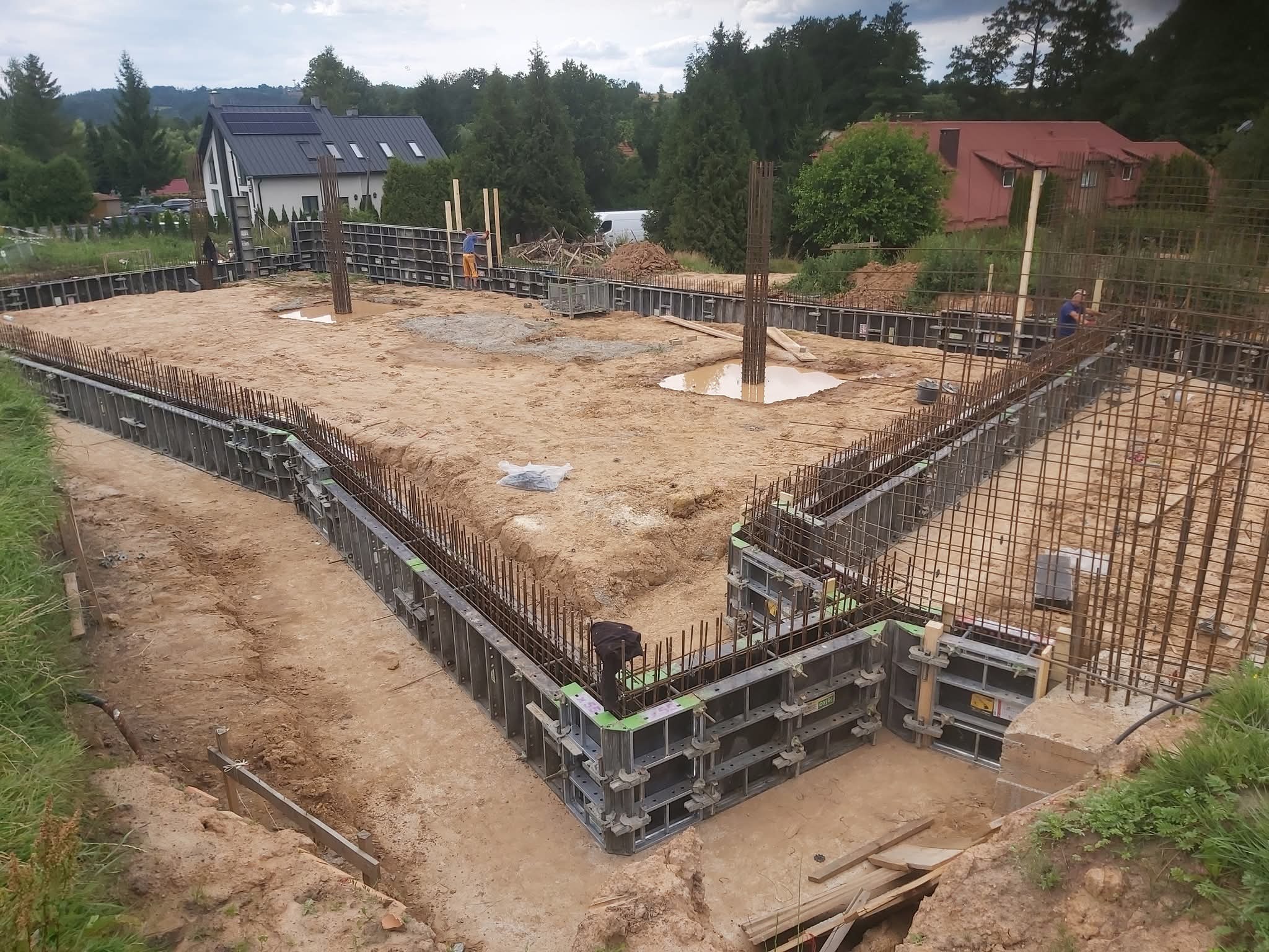 Zbrojenie fundamentów pod dom jednorodzinny z użyciem szalunków systemowych. Widoczne wystające pręty zbrojeniowe i pracownicy budowlani w tle.
