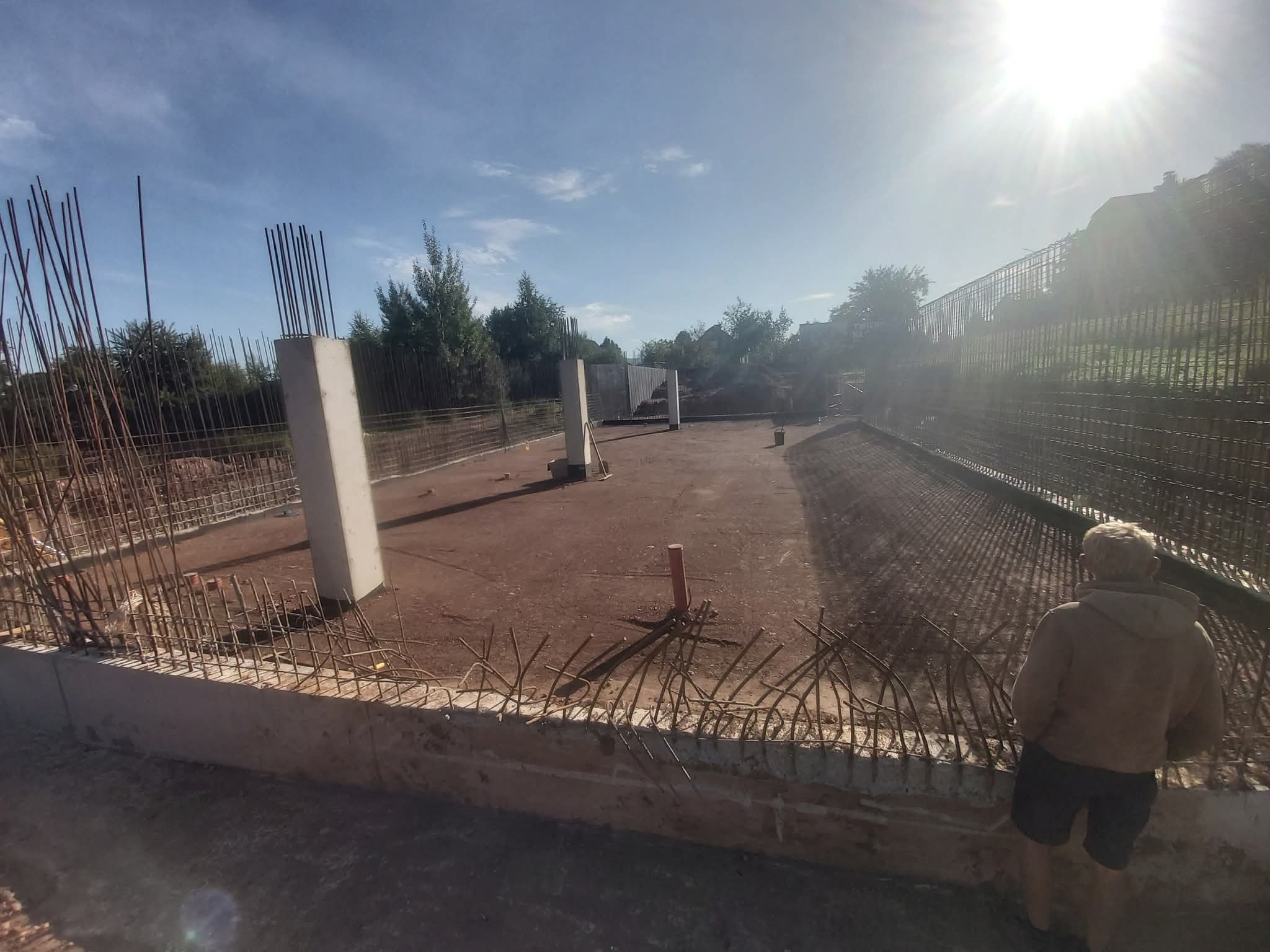 Zbrojenie fundamentów pod budowę domu jednorodzinnego w słoneczny dzień. Widoczne wystające pręty zbrojeniowe i betonowe słupy. Osoba obserwuje postęp prac.