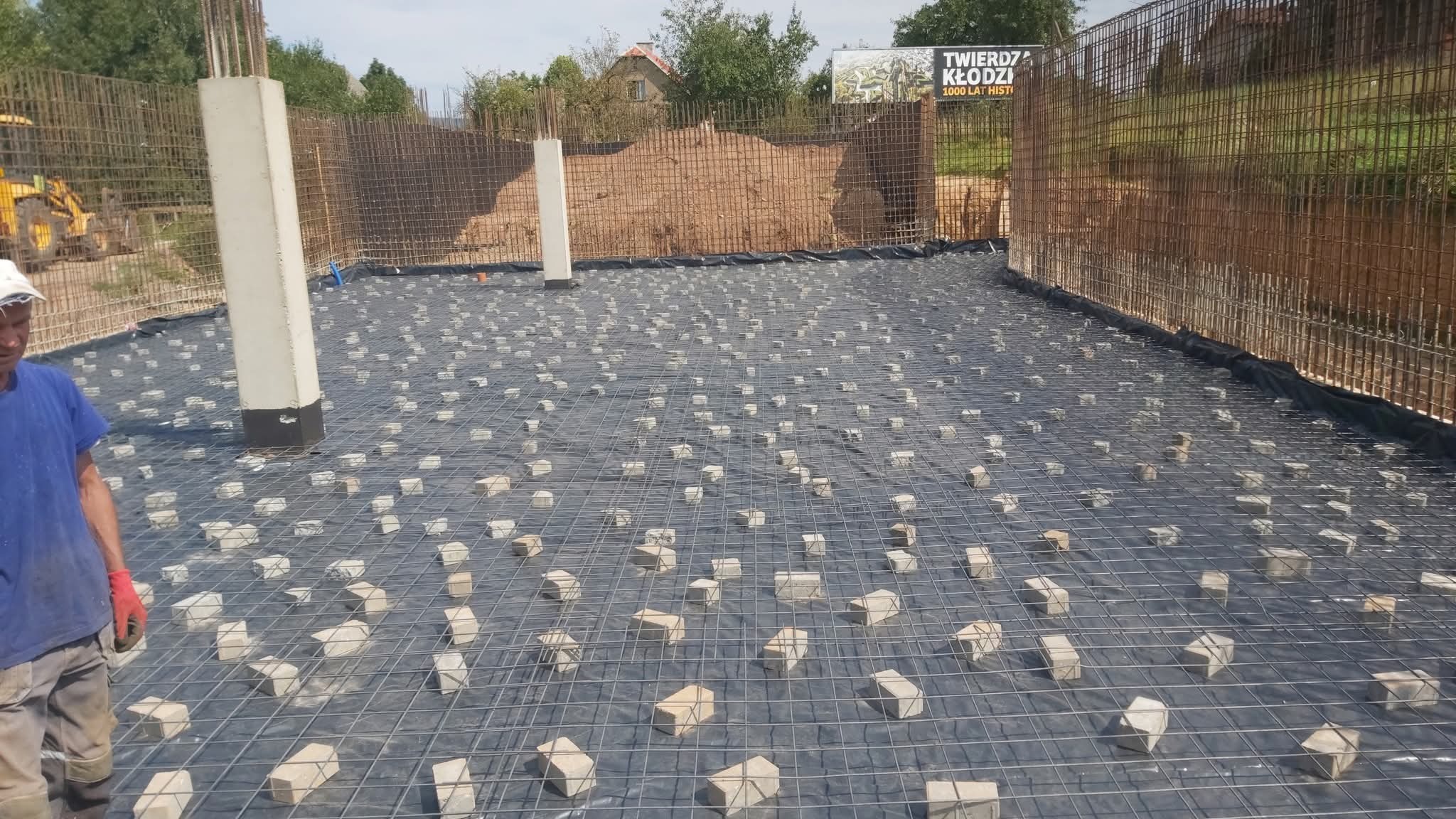 Zbrojenie fundamentu z siatki stalowej na czarnej folii, podparte kostkami. W tle pionowe zbrojenie ścian fundamentowych i częściowo widoczny pracownik w czerwonej rękawicy.