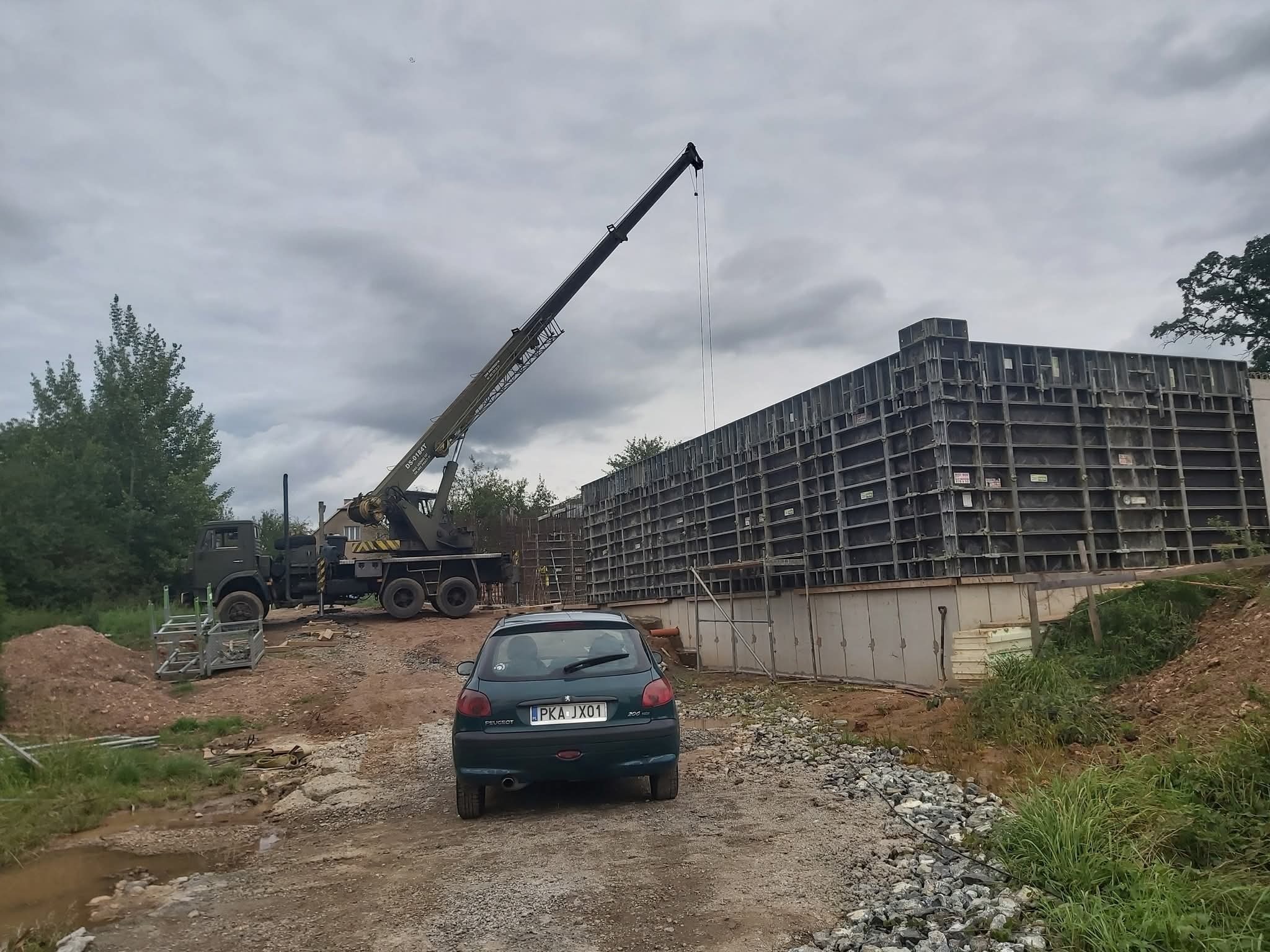 Budowa fundamentów: szalunek zbrojeniowy, żuraw budowlany i zielony Peugeot 206 na placu budowy w pochmurny dzień. Widok z tyłu samochodu.