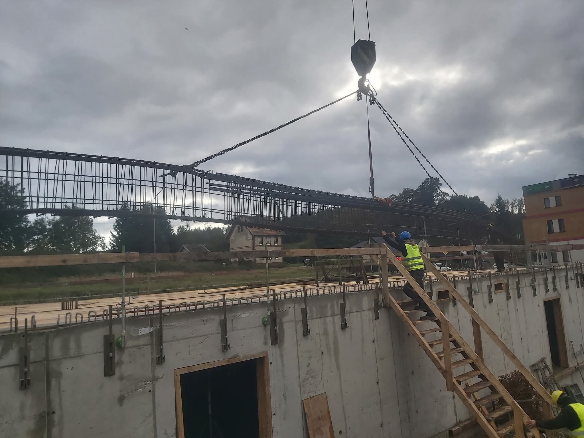 Montaż zbrojenia fundamentów za pomocą dźwigu na budowie. Widoczni pracownicy w kaskach i kamizelkach. Szalunki betonowe z widocznymi otworami.
