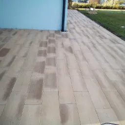 Ułożona kostka brukowa imitująca drewniane deski na tarasie przy domu, widoczny fragment ściany budynku, rynna spustowa, trawnik i elementy systemu nawadniania.