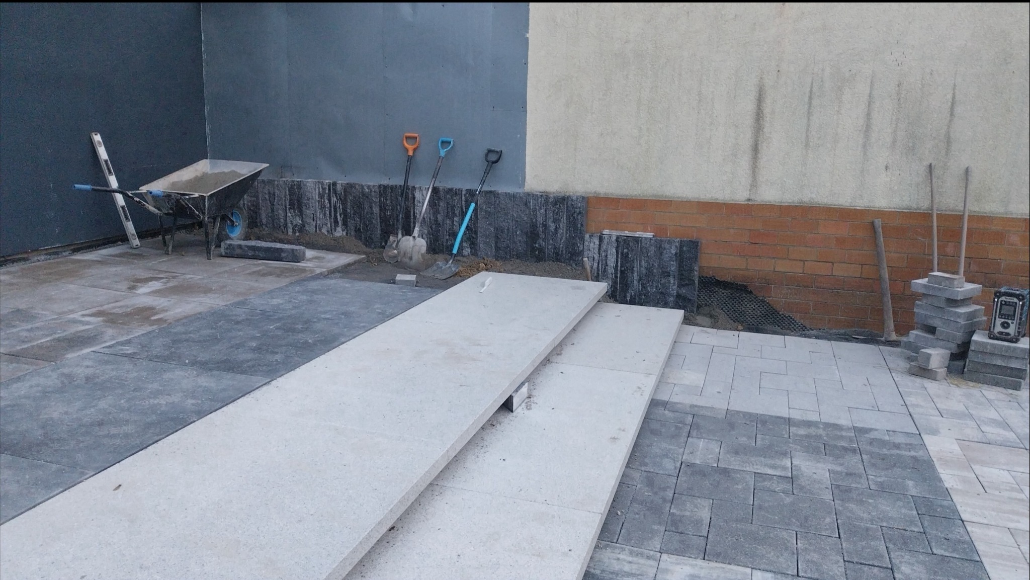 Etap prac brukarskich w Leśnicy: ułożone płyty tarasowe, przygotowane podłoże pod kostkę, narzędzia pracy. Widoczny wózek z zaprawą i poziomica.