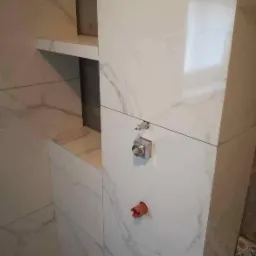 Białe płytki imitujące marmur z widocznymi instalacjami hydraulicznymi i wnęką na półki, prawdopodobnie w trakcie prac wykończeniowych w łazience w Poznaniu.