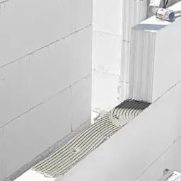 Ściana z białych bloczków z betonu komórkowego w trakcie murowania, widoczna warstwa kleju z charakterystycznym wzorem nałożona na spoinę poziomą, obok leży gumowy młotek.