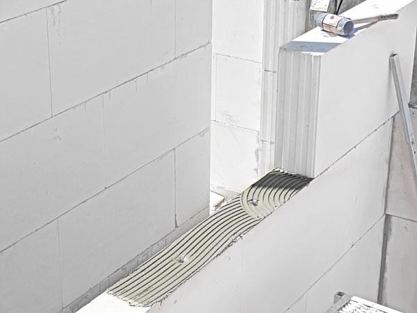 Ściana z białych bloczków z betonu komórkowego w trakcie murowania, widoczna warstwa kleju z charakterystycznym wzorem nałożona na spoinę poziomą, obok leży gumowy młotek.