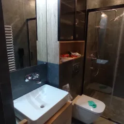 Nowoczesna łazienka z umywalką nablatową na drewnianej szafce, wiszącą toaletą z zieloną ściereczką, kabiną prysznicową i szafką z czarnymi ramami, widoczne akcesoria montażowe.