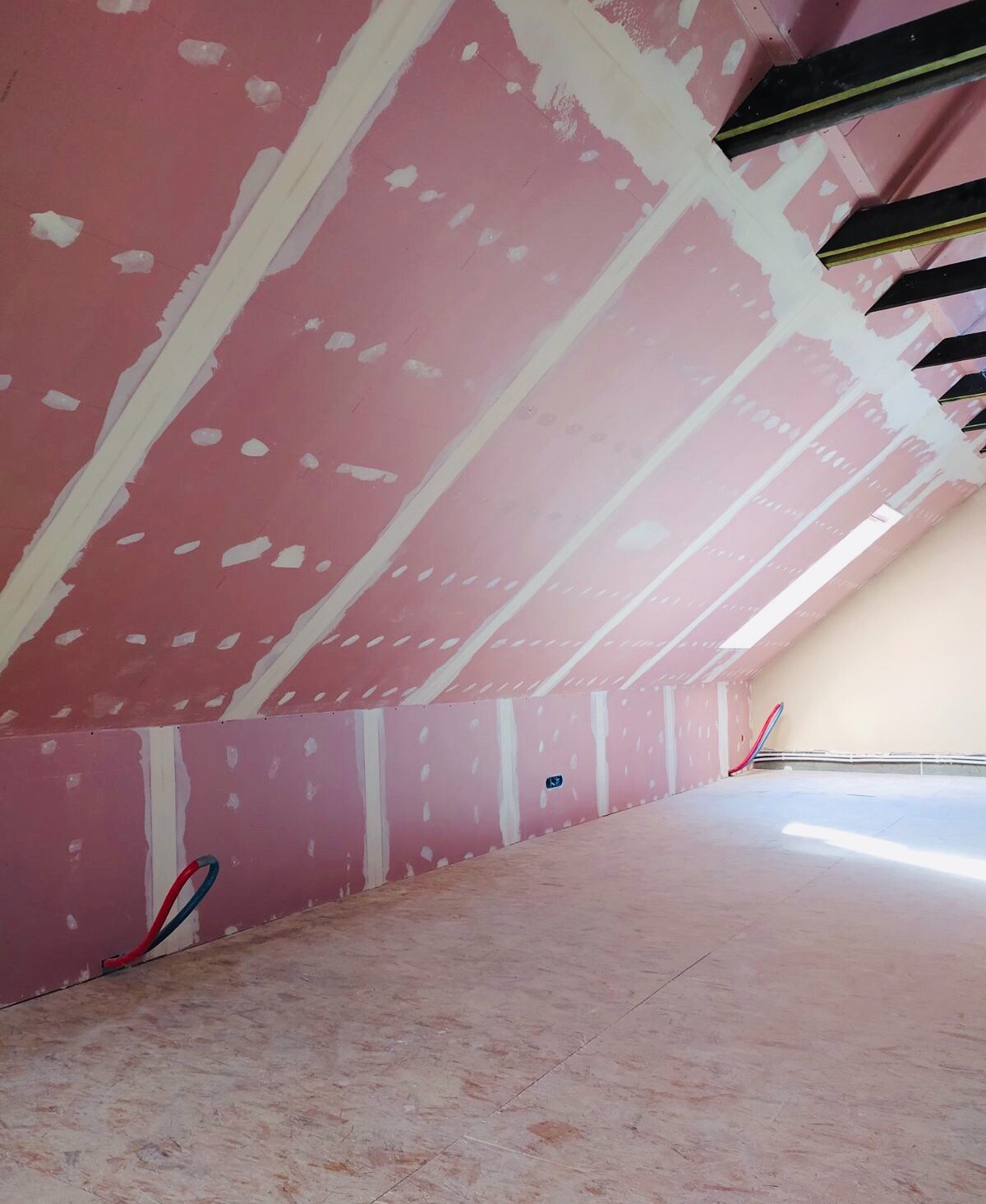 Poddasze w trakcie wykańczania: różowe płyty karton-gips na skośnym suficie i ścianie, łączenia zaszpachlowane na biało, widoczne fragmenty instalacji elektrycznej i rury grzewcze, surowa podłoga...