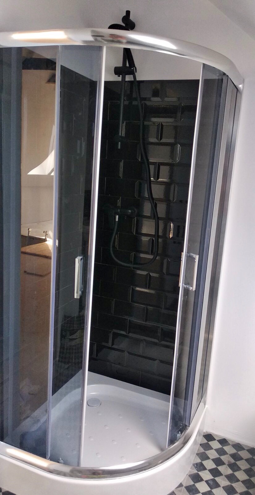 Narożna kabina prysznicowa z czarnymi, błyszczącymi płytkami ułożonymi w cegiełkę i czarną armaturą, widoczna szachownica na podłodze.