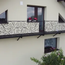 Elegancki balkon z czarną, kutą balustradą o dekoracyjnym, spiralnym wzorze, zamontowany przy beżowej elewacji domu z brązową stolarką okienną i kwiatami w donicach.