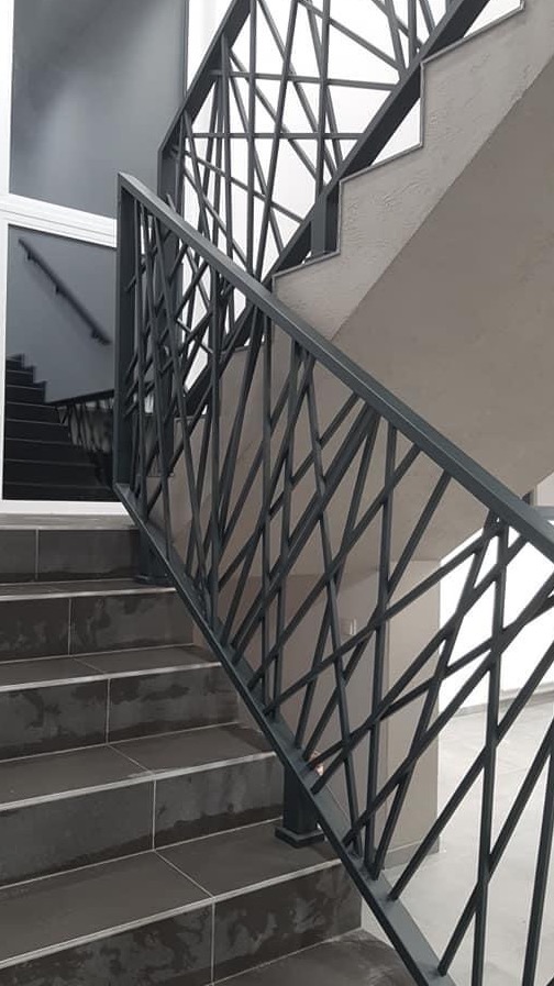 Schody z ciemnymi płytkami i nowoczesną, geometryczną balustradą w kolorze grafitowym. Widoczne okno z kolejnymi schodami w tle.
