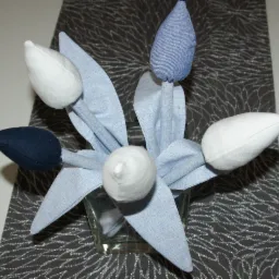 Dekoracyjny bukiet tulipanów uszytych z tkaniny w odcieniach bieli, błękitu i granatu, umieszczony w szklanym wazonie na tle ciemnoszarej serwety ze srebrnym wzorem.