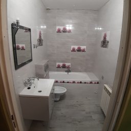 M.S KaFelki - Wykończona łazienka z wanną obudowaną płytkami z motywem kwiatowym, umywalką z szafką, lustrem w czarnej ramie i grzejnikiem na ścianie, widok zza futryny drzwi.