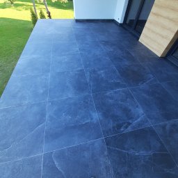 M.S KaFelki - Wyłożony ciemnymi płytkami taras z widokiem na trawnik, widoczne buty wykonawcy.