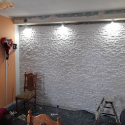 M.S KaFelki - Ściana wykończona dekoracyjną białą cegłą, oświetlona trzema punktowymi lampami, widoczny fragment drabiny i krzesło w remonowanym pomieszczeniu.