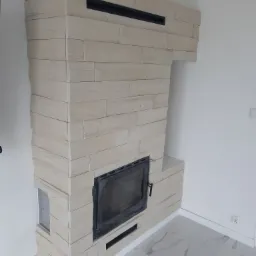 Obudowa kominka wykończona płytkami imitującymi drewno, z czarną wkładką kominkową i marmurową podłogą.