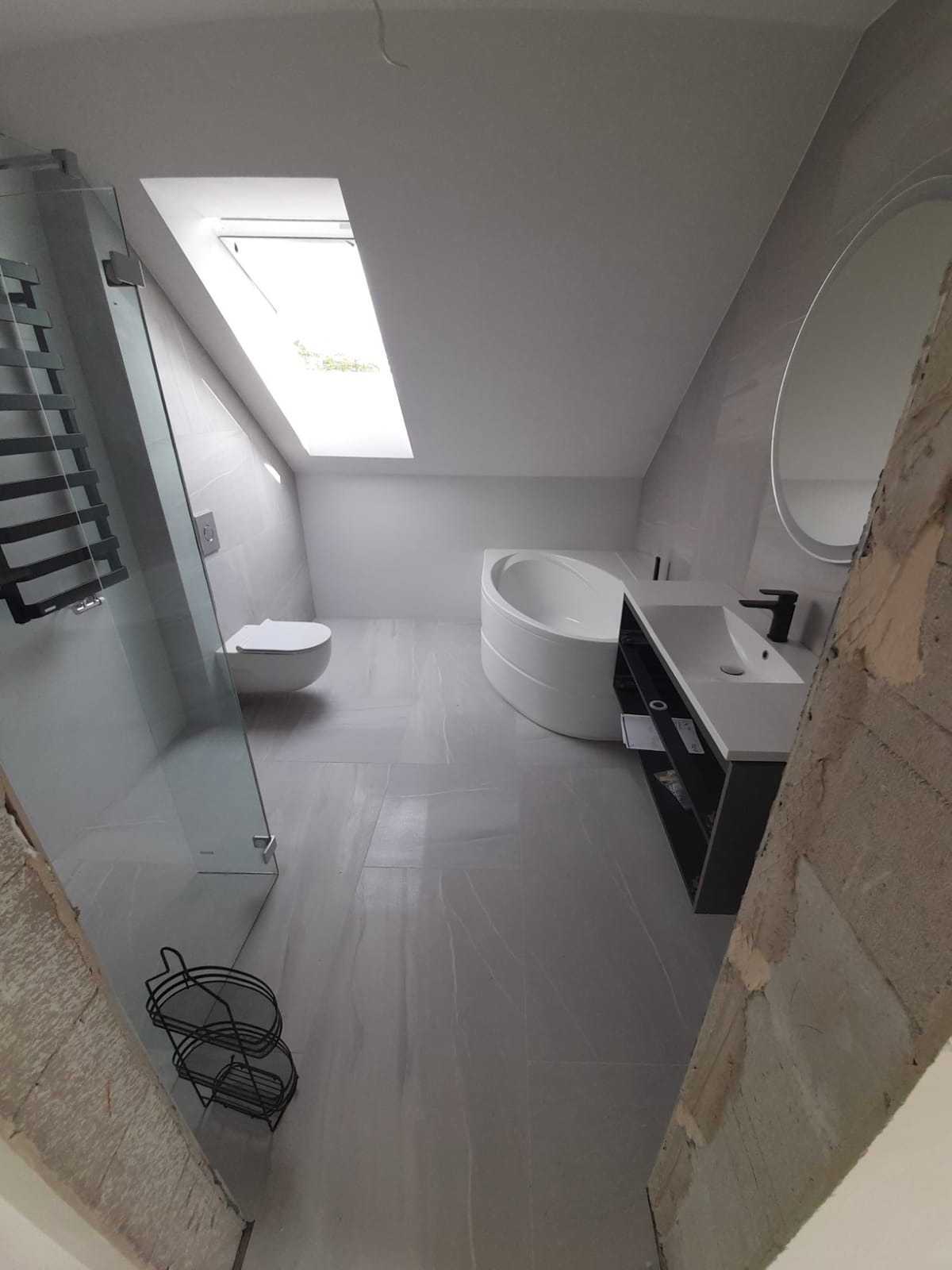 Nowoczesna łazienka na poddaszu z szarymi płytkami, kabiną prysznicową ze szklanymi drzwiami, owalną wanną i umywalką z szafką, oświetlona przez okno dachowe.