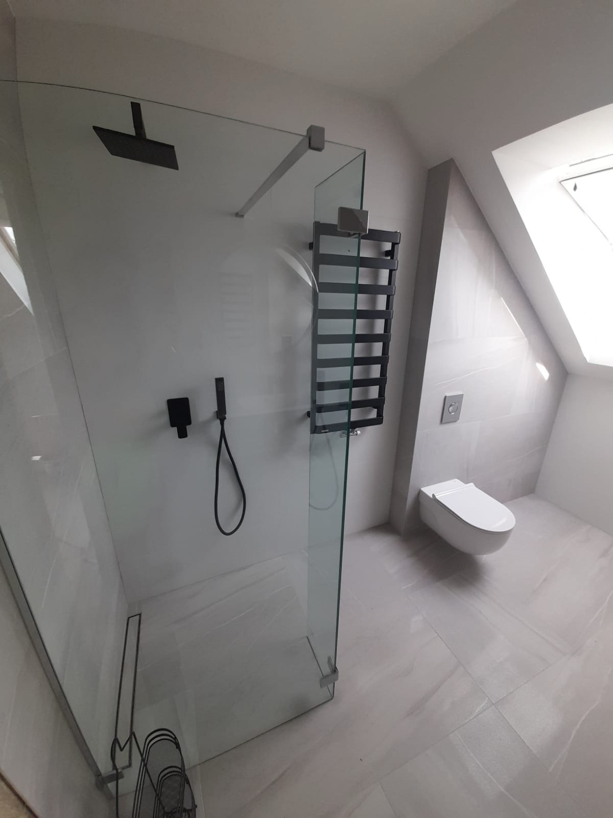 Nowoczesna łazienka na poddaszu z kabiną prysznicową typu walk-in, podwieszaną toaletą i grzejnikiem drabinkowym w kolorze grafitowym.