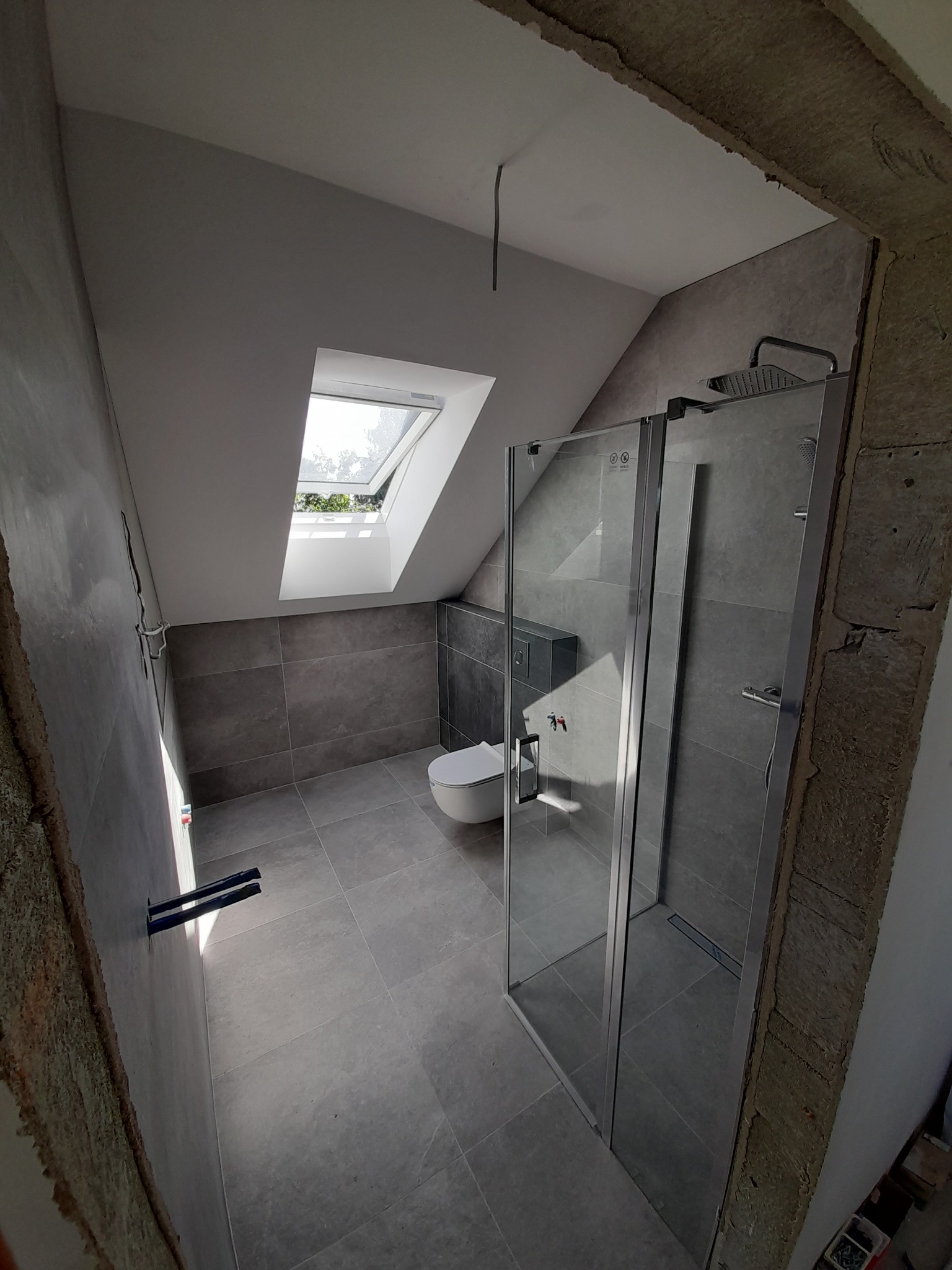 Szara łazienka na poddaszu z oknem dachowym, kabiną prysznicową typu walk-in i wiszącą toaletą, widoczne niedokończone wykończenia ścian.