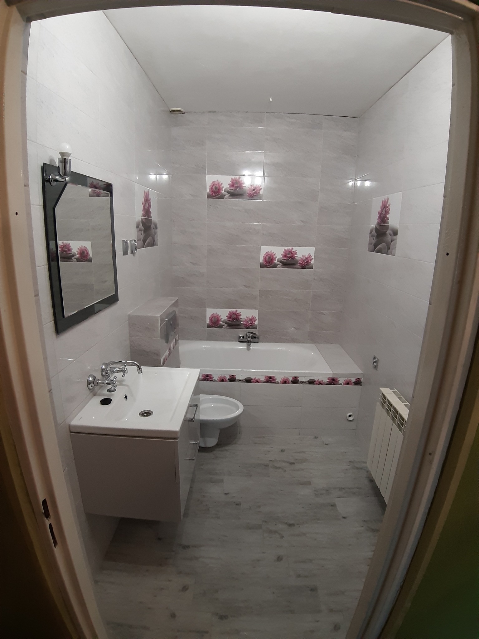 Wykończona łazienka z wanną obudowaną płytkami z motywem kwiatowym, umywalką z szafką, lustrem w czarnej ramie i grzejnikiem na ścianie, widok zza futryny drzwi.