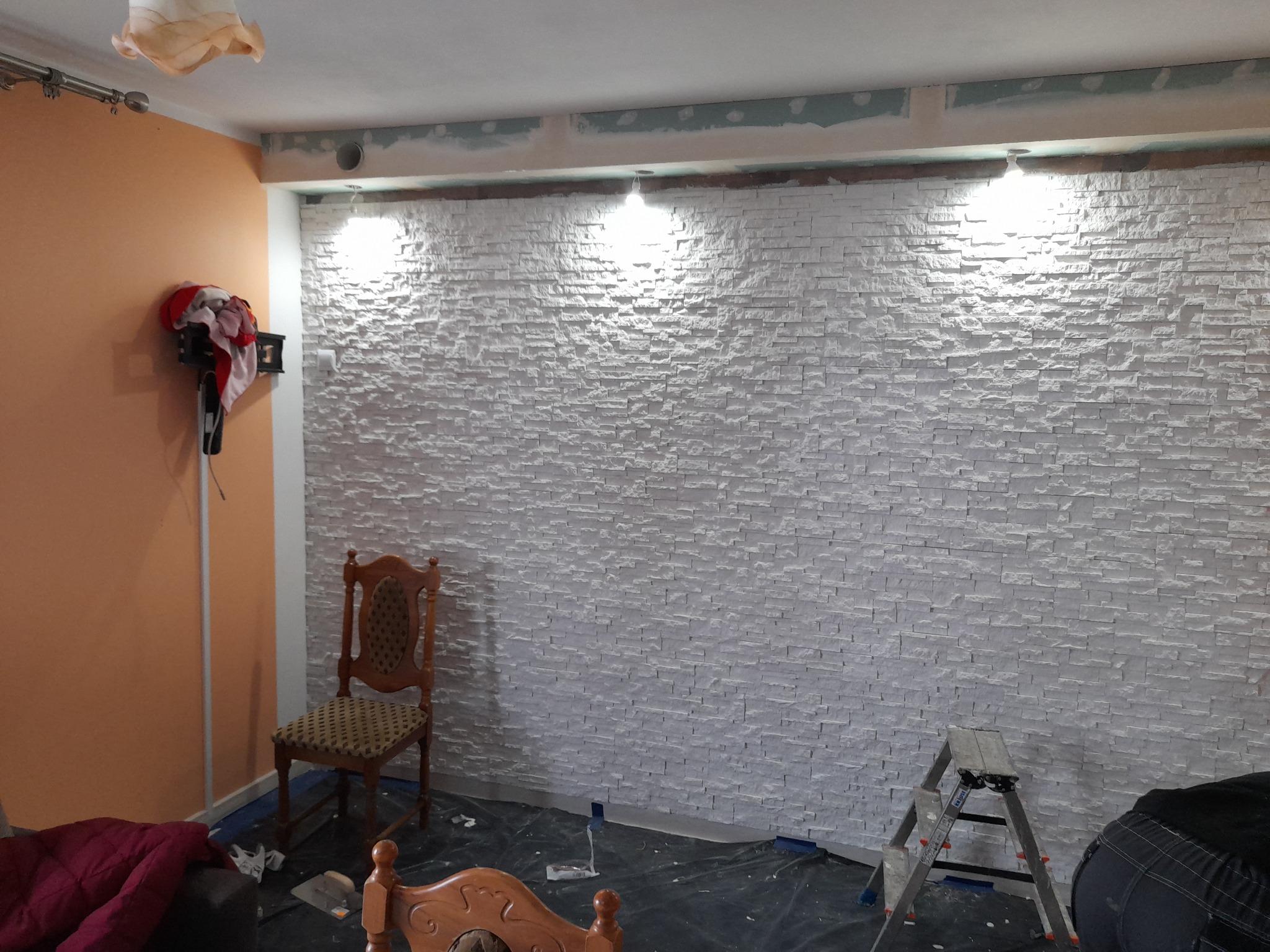 Ściana wykończona dekoracyjną białą cegłą, oświetlona trzema punktowymi lampami, widoczny fragment drabiny i krzesło w remonowanym pomieszczeniu.