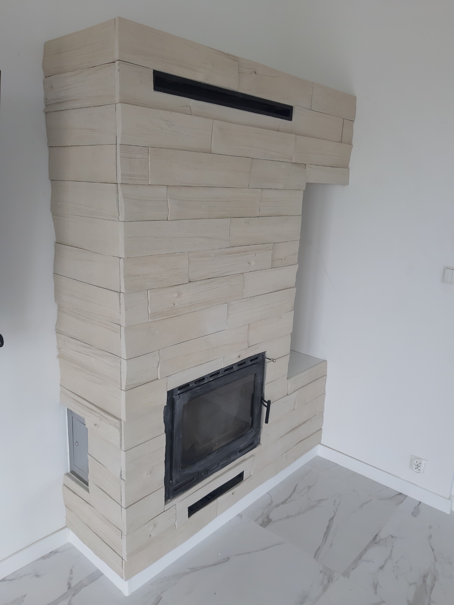 Obudowa kominka wykończona płytkami imitującymi drewno, z czarną wkładką kominkową i marmurową podłogą.