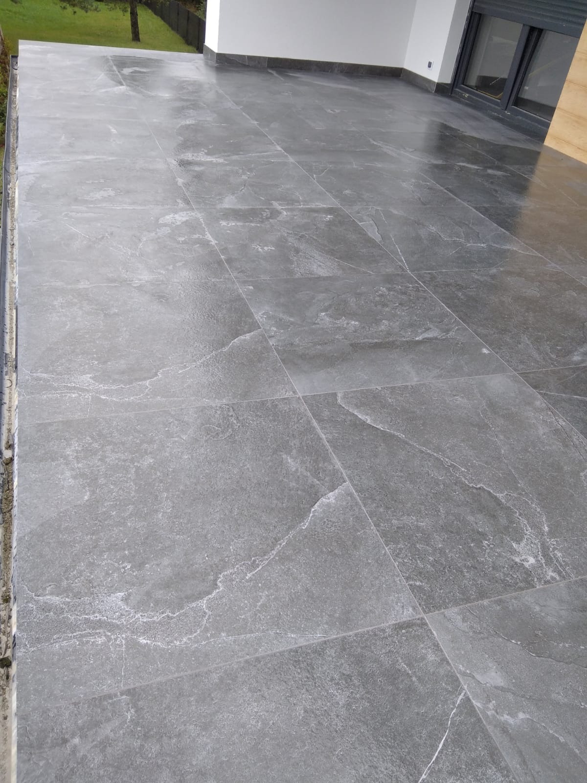 Ułożona szara, kamieniopodobna terakota na podłodze balkonu, widoczne fugi i fragment ściany z oknem.