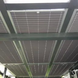 Zewnętrzne osłony przeciwsłoneczne w kolorze szarym, zamontowane pod drewnianą konstrukcją pergoli, widok od dołu.