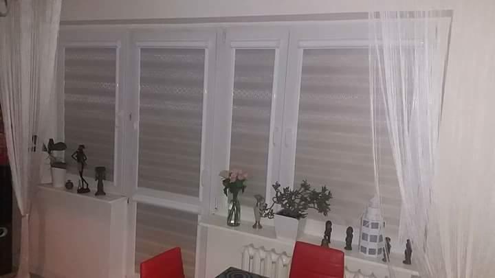 Trzy okna z białymi ramami, zasłonięte poziomymi osłonami okiennymi, z dekoracjami na parapecie i firankami po bokach.