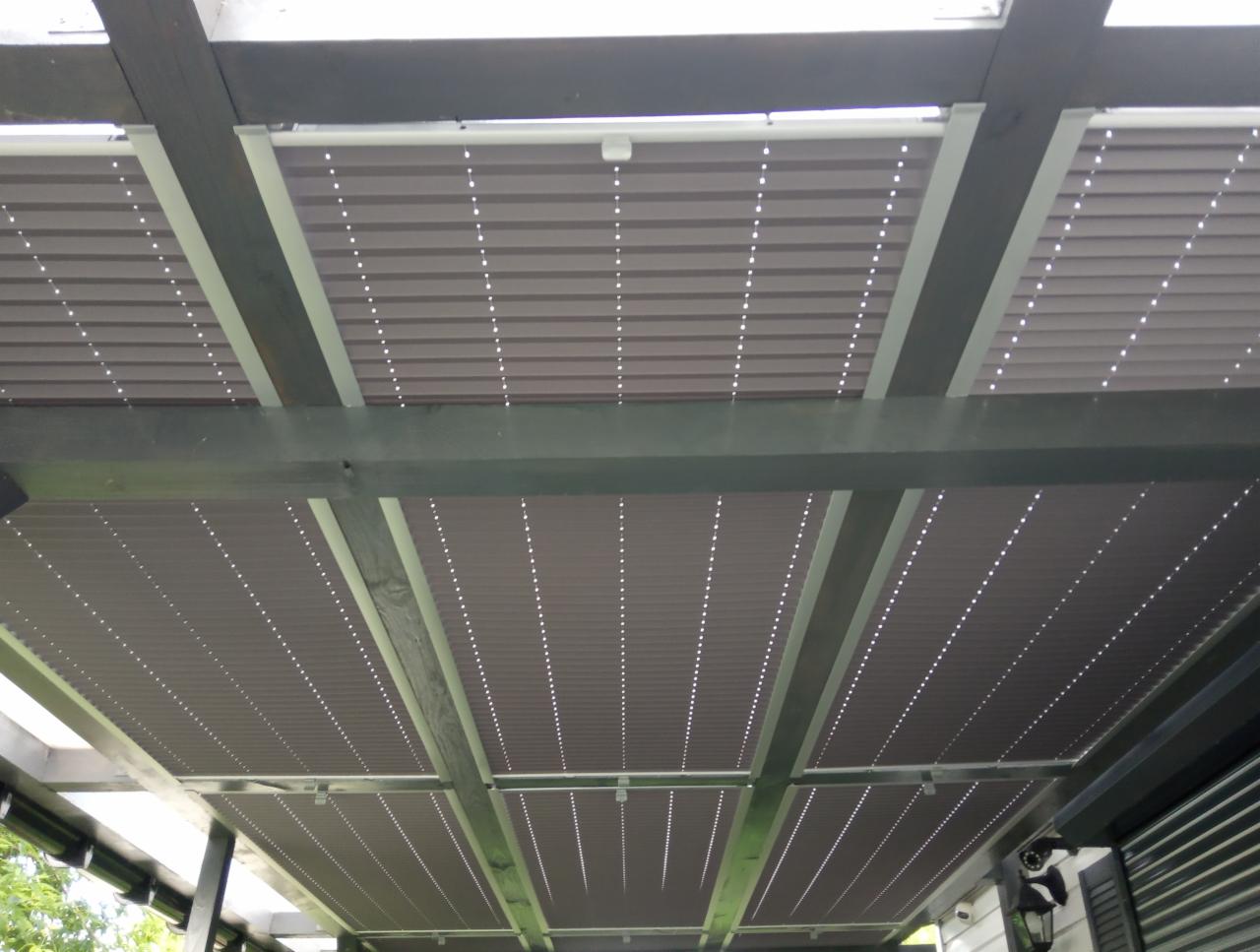 Zewnętrzne osłony przeciwsłoneczne w kolorze szarym, zamontowane pod drewnianą konstrukcją pergoli, widok od dołu.