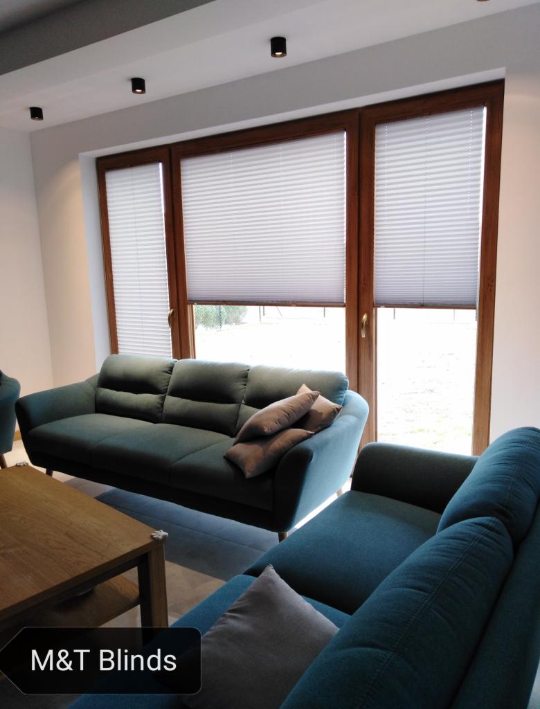 Salon z drewnianą stolarką okienną i białymi plisowanymi osłonami okiennymi, widoczny fragment turkusowej sofy i stolika kawowego.