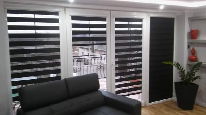 Wnętrze pokoju z dużym oknem balkonowym, na którym zamontowano czarne i białe pasiaste osłony okienne typu dzień i noc, częściowo zasłonięte, z widokiem na balkon i budynki na zewnątrz.